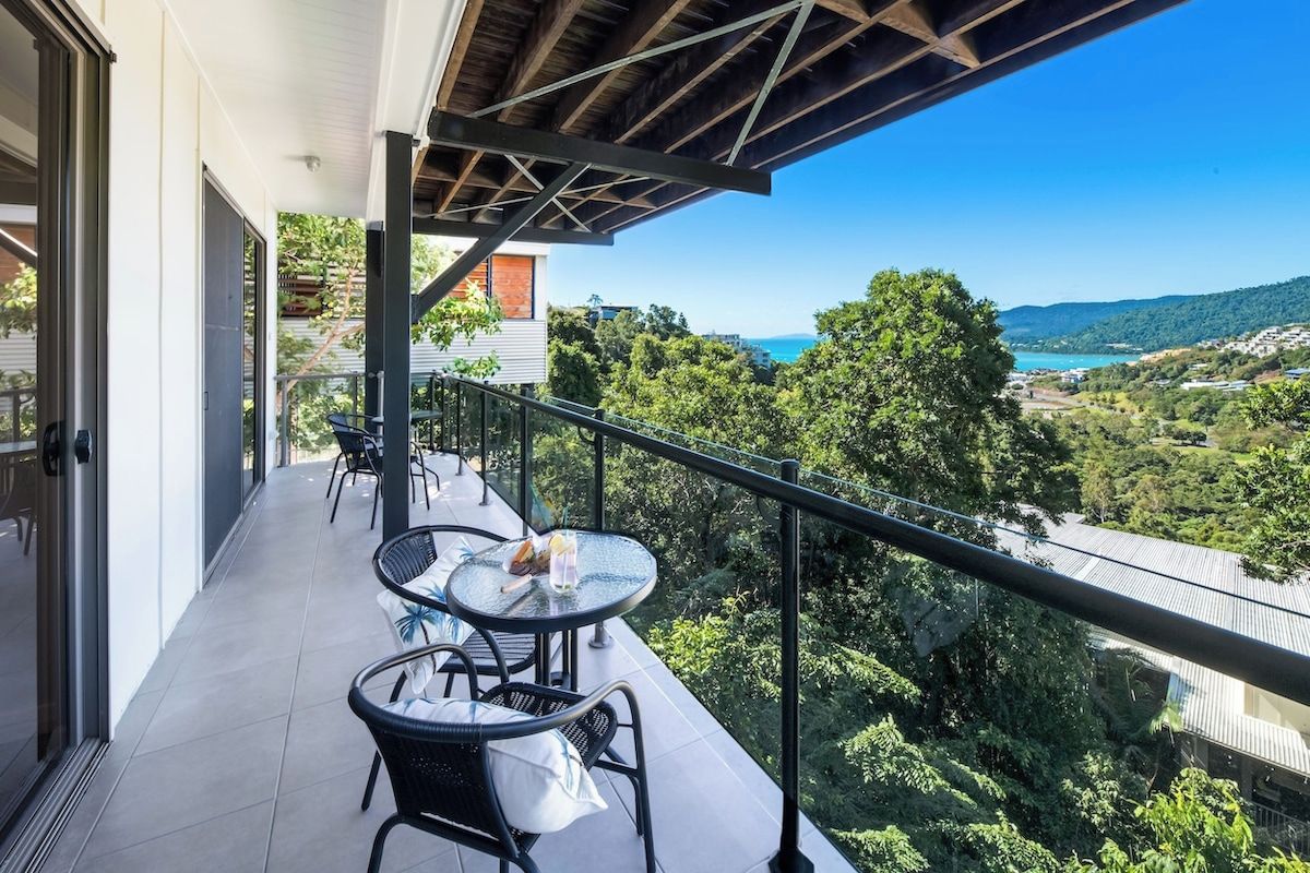 A balcony with outdoor seating, a view of a coastal landscape, and a building with a modern architectural style.