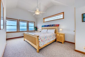 A bedroom with a bed, bedside tables, a ceiling fan, and large windows overlooking the sea.