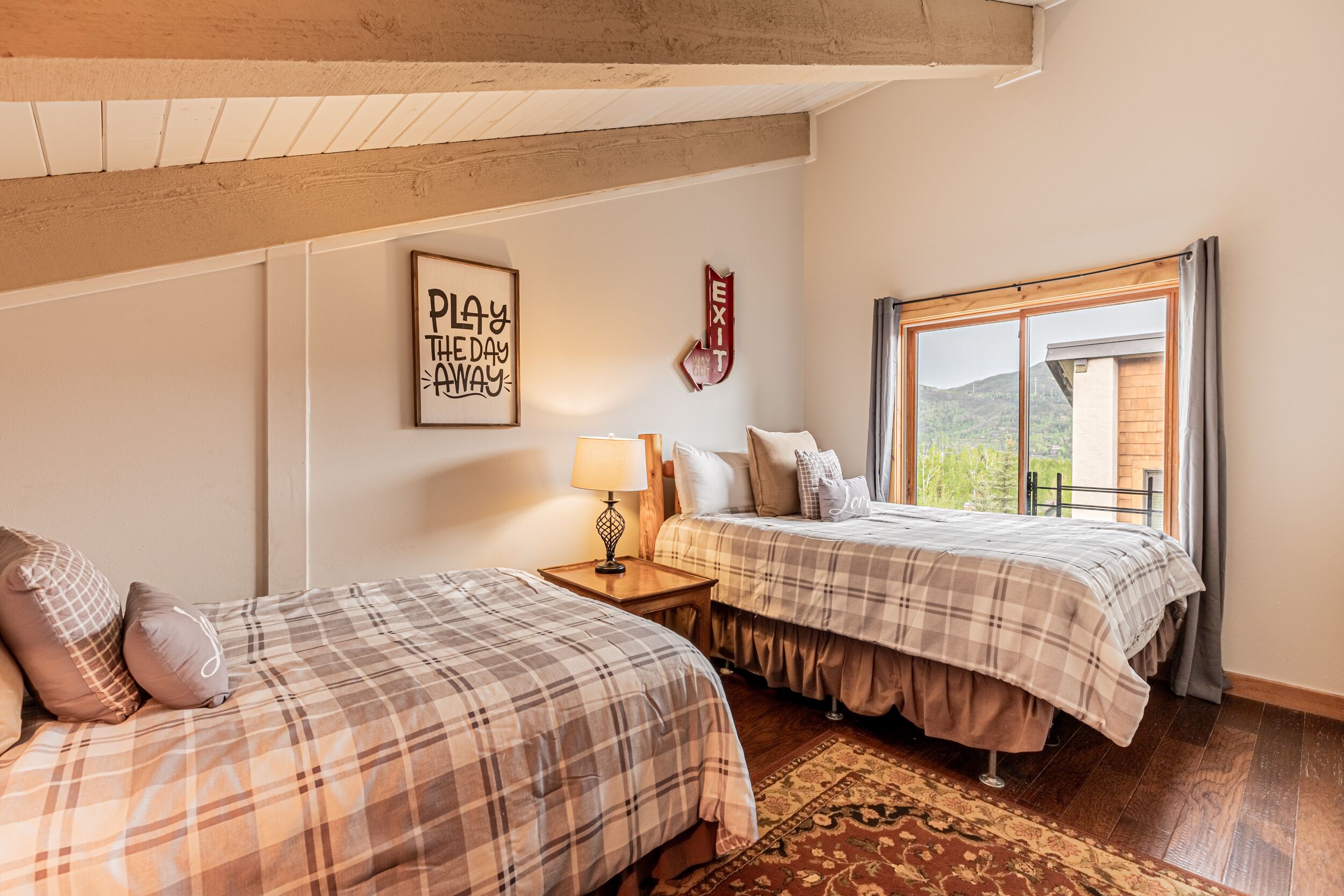 A bedroom with two beds, a nightstand, a lamp, a framed quote on the wall, and a window with a view of the outdoors.