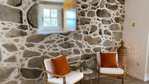 Une pièce aux murs de pierre, deux chaises en bois avec des coussins, une petite table ronde et une fenêtre donnant sur un paysage verdoyant.