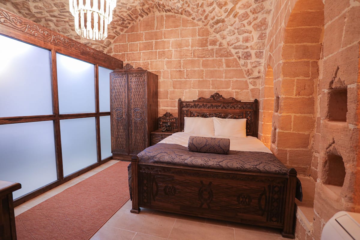 A bedroom with a stone wall, a wooden bed with intricate carvings, a large mirror, and a chandelier.
