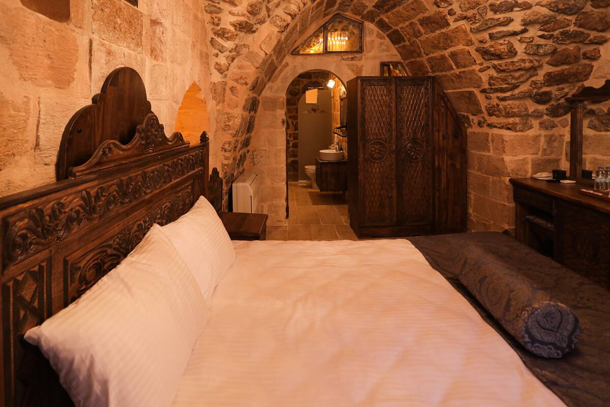 A bedroom with a stone wall, a wooden bed with a headboard, a wooden bench, and a small table with a mirror and toiletries.