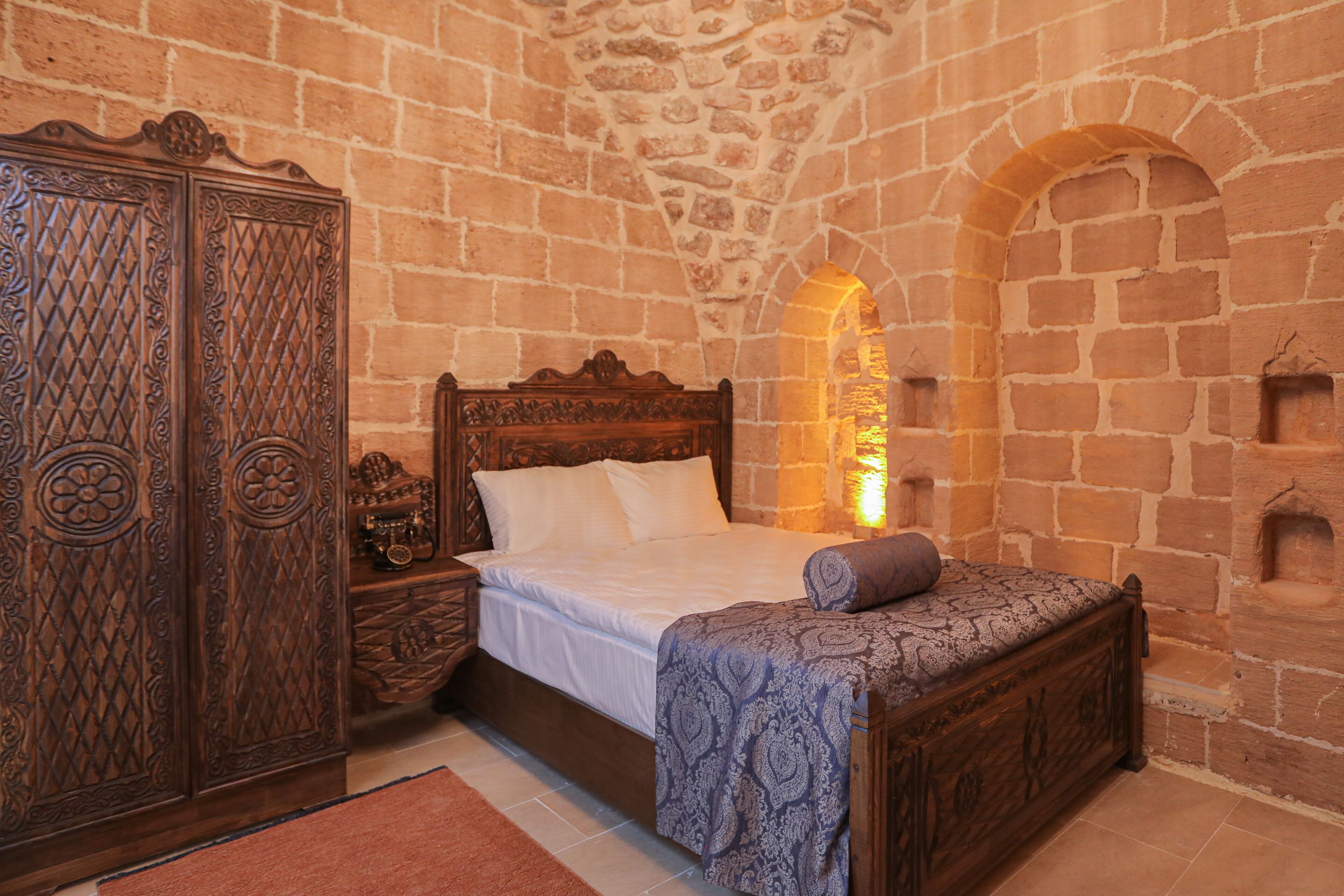 Un dormitorio con cama de madera, una mesita de noche y un armario. La habitación tiene paredes de piedra y un ventanal de arco.