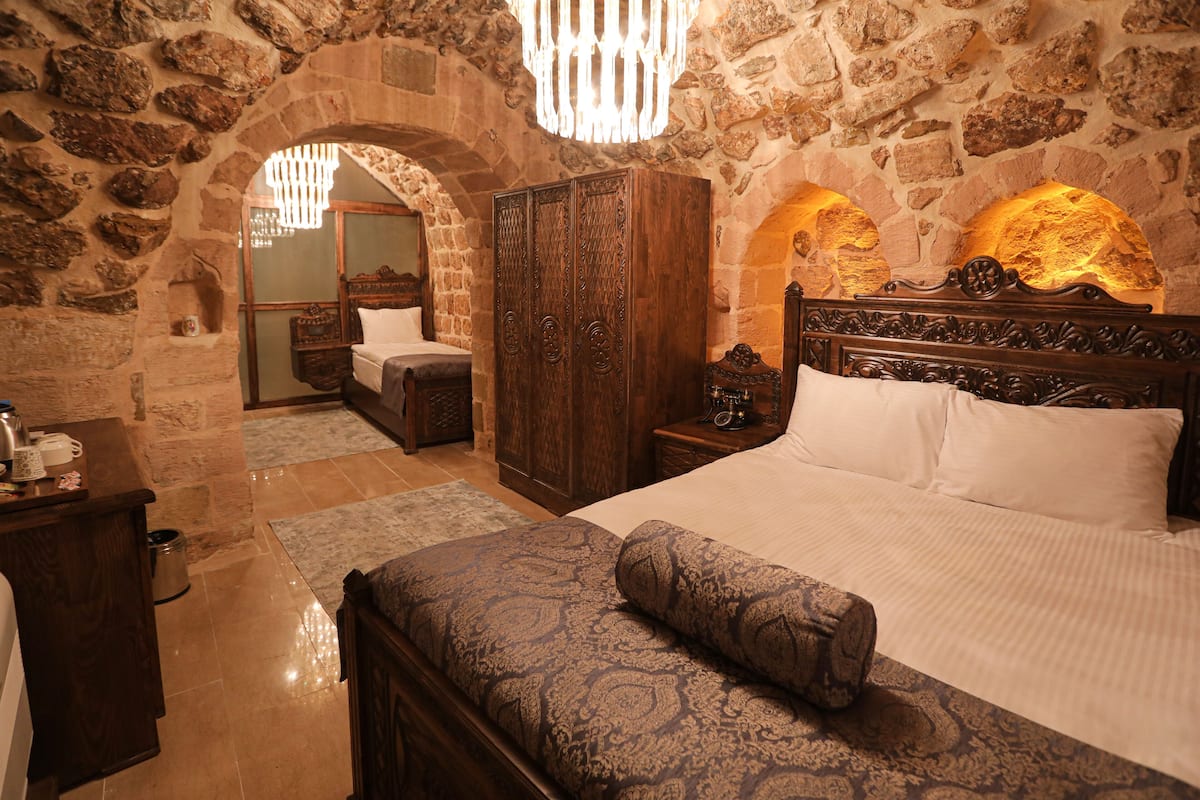 A bedroom with a stone wall, a wooden wardrobe, a bed with white linens, and a patterned bench.
