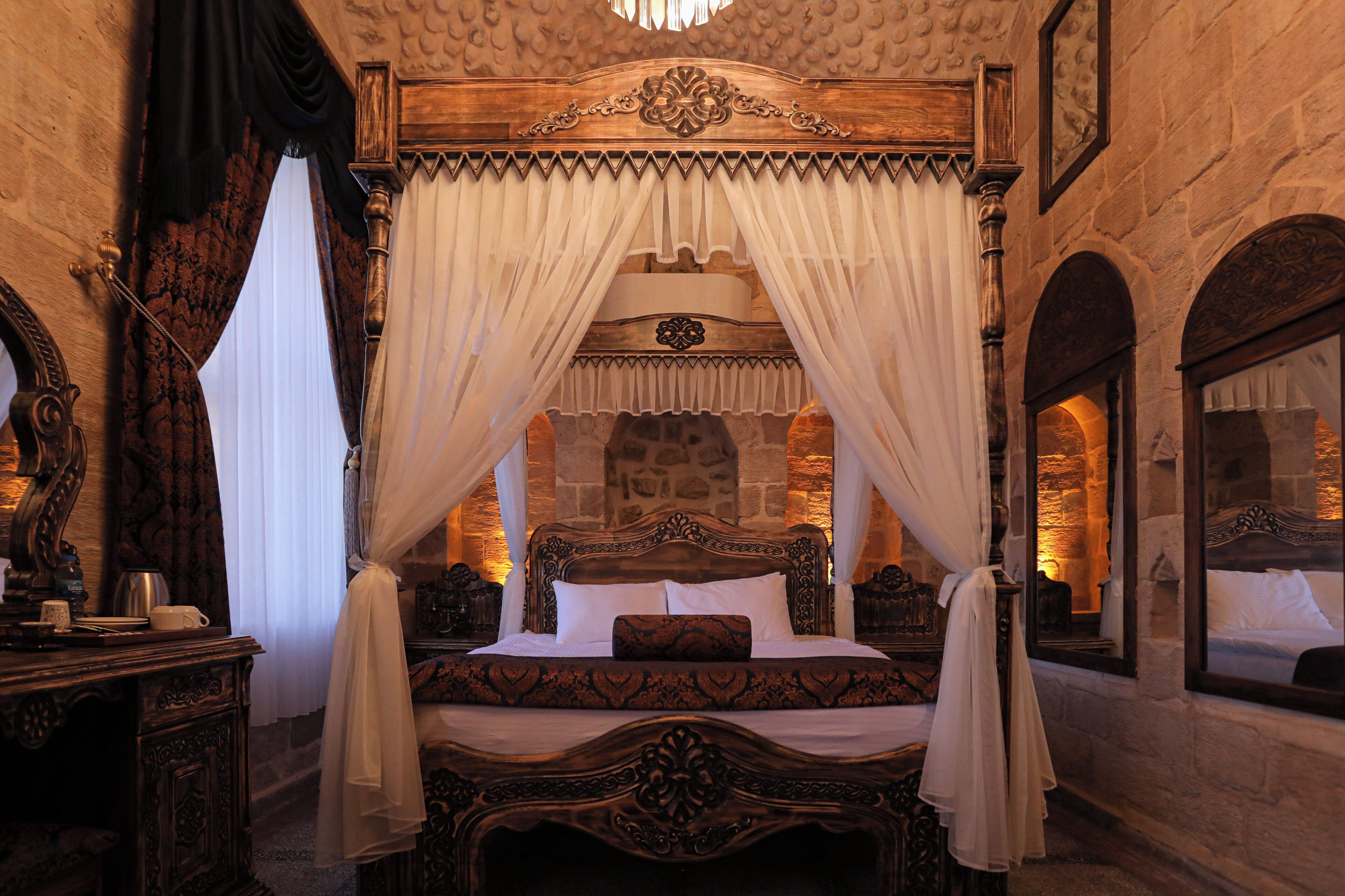 A four-poster canopy bed with a stone wall backdrop, a wooden dresser with a mirror, and a small table with a cup and saucer.