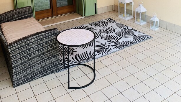 Un balcon agrémenté d’un canapé en osier, d’une petite table ronde, d’un tapis à motifs et de lanternes blanches.