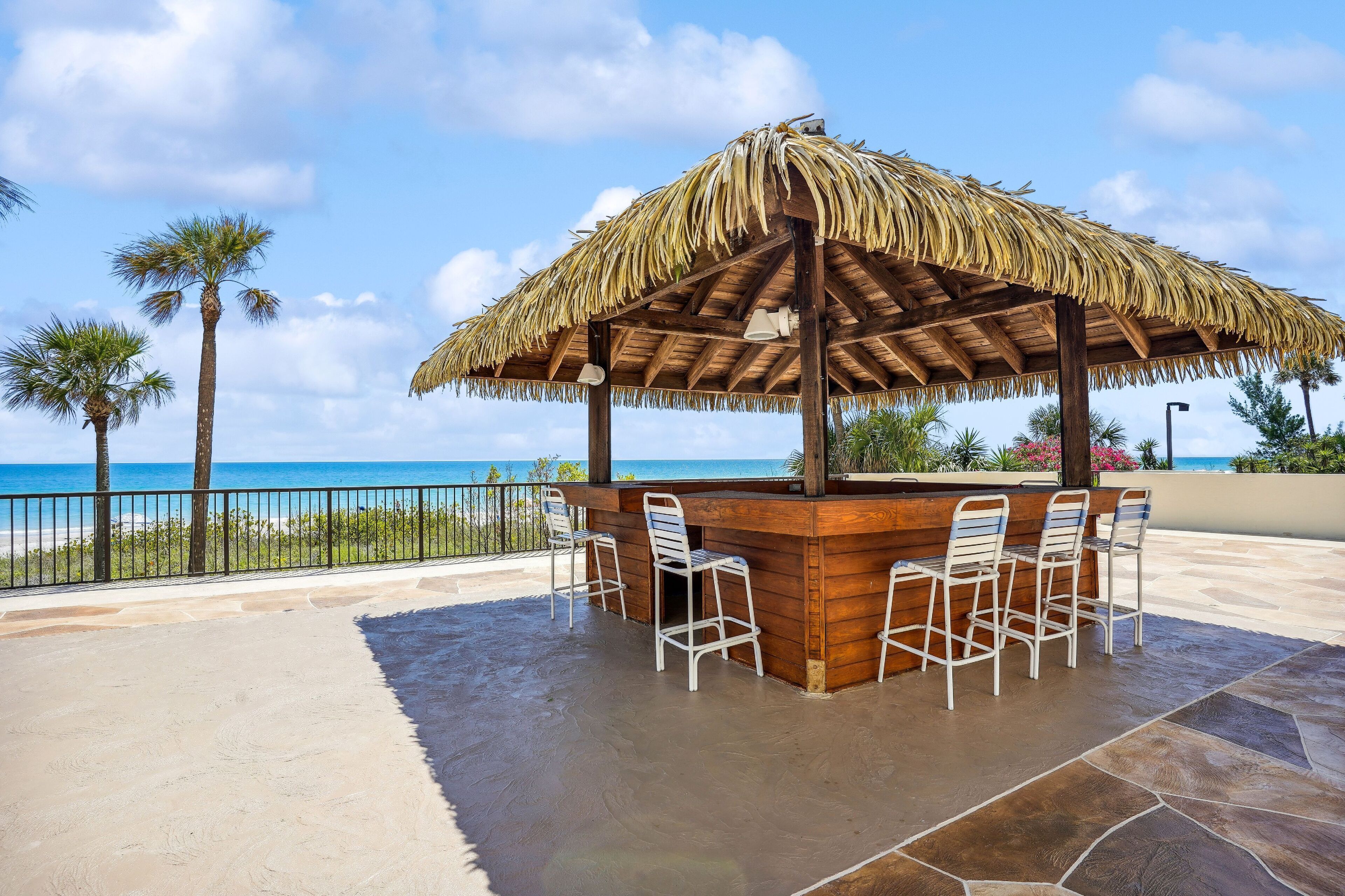 An outdoor bar area with a thatched roof, stone flooring, and white metal chairs.