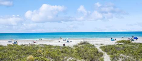 Een strand met helderblauw water, een zandstrand en schaarse begroeiing.