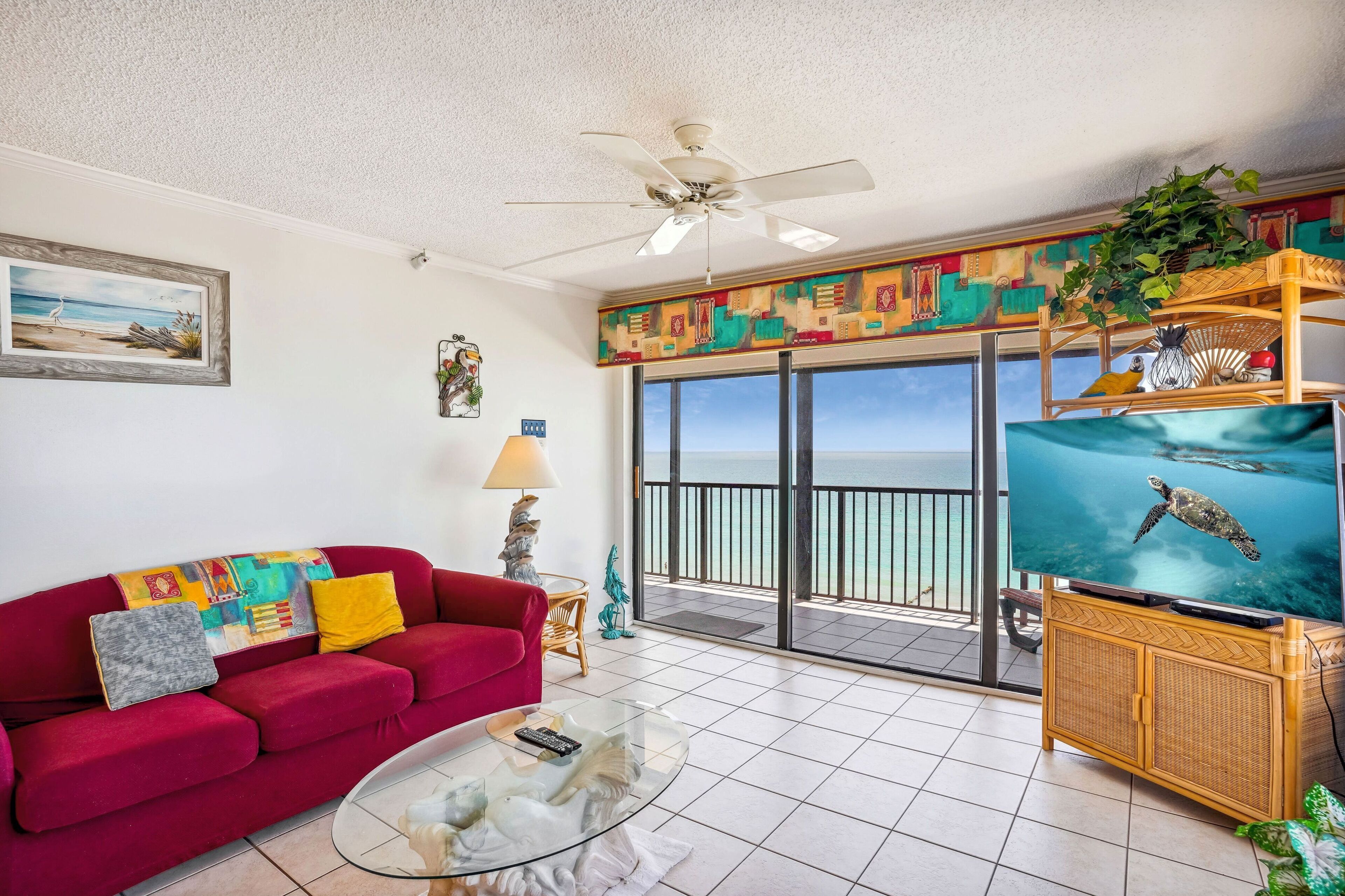 A living room with a red sofa, a glass coffee table, a ceiling fan, a TV, and a balcony with a view of the ocean.