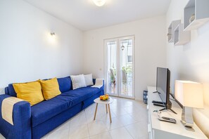 Un salon moderne avec un canapé bleu, une petite table ronde sur laquelle sont disposés des fruits, un téléviseur à écran plat et une lampe.