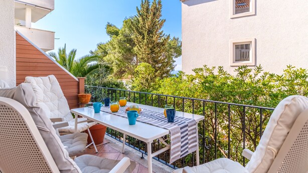 En balkon med et bord dækket til to, udendørs siddepladser og udsigt til en bygning og træer.