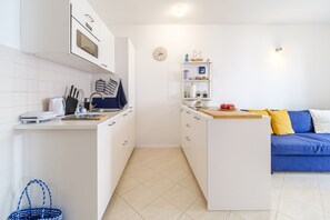 Une cuisine compacte avec des meubles blancs, un four à micro-ondes et un petit îlot central doté d’une planche à découper. Le salon adjacent est équipé d’un canapé bleu agrémenté de coussins jaunes.