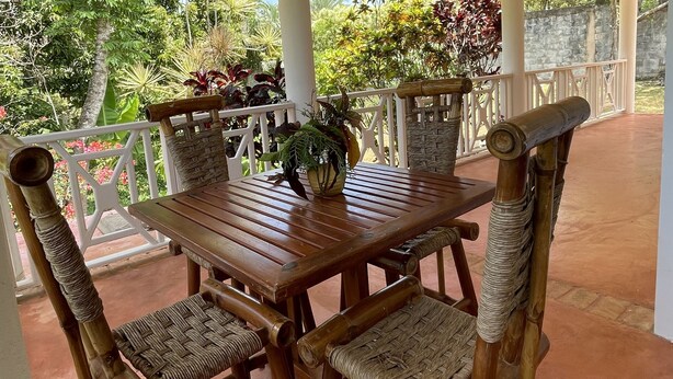A wooden table and chairs with woven seats, a potted plant, and a view of lush greenery and a stone wall. - Casa yann  (Las Galeras)
