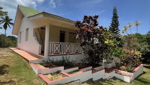 A single-story house with a covered porch, surrounded by a well-maintained garden with various plants and a palm tree. - Casa yann  (Las Galeras)