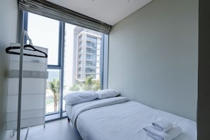 A bedroom with a bed, a hanging clothes rack, and a large window with a view of a building and palm trees.