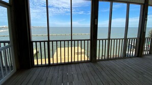 A covered wooden deck with large windows overlooking a beach and ocean.