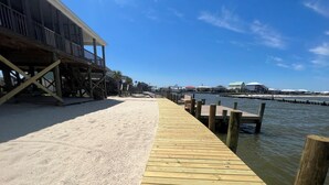 A wooden pier extending into calm water, with a sandy beach on one side and a building with a balcony on the other.