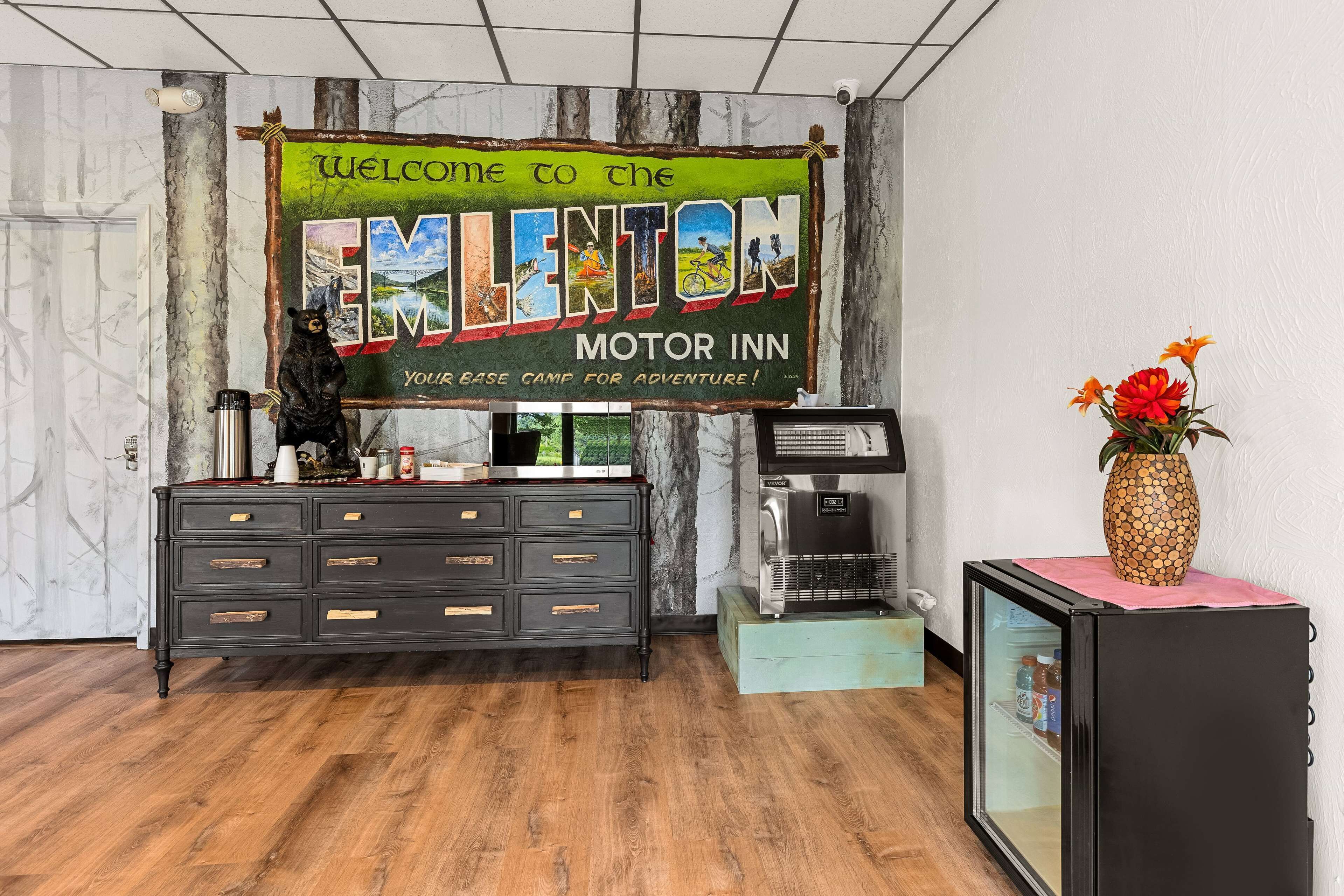 Una zona de recepción con piso de madera, un aparador negro con cajones, una cafetera, un refrigerador y un cartel de bienvenida para el EMILTON MOTOR INN.