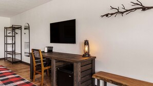 Ein kleiner Raum mit einem Holztisch, einem Stuhl, einer Bank, einem kleinen schwarzen Kühlschrank, einem wandmontierten Fernseher, einem Kleiderhaken und einem dekorativen Zweig an der Wand.