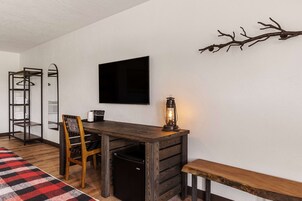 Quarto compacto com uma escrivaninha de madeira, uma cadeira, um banco, um refrigerador preto pequeno, uma TV fixada na parede, um cabideiro e um galho decorativo na parede.