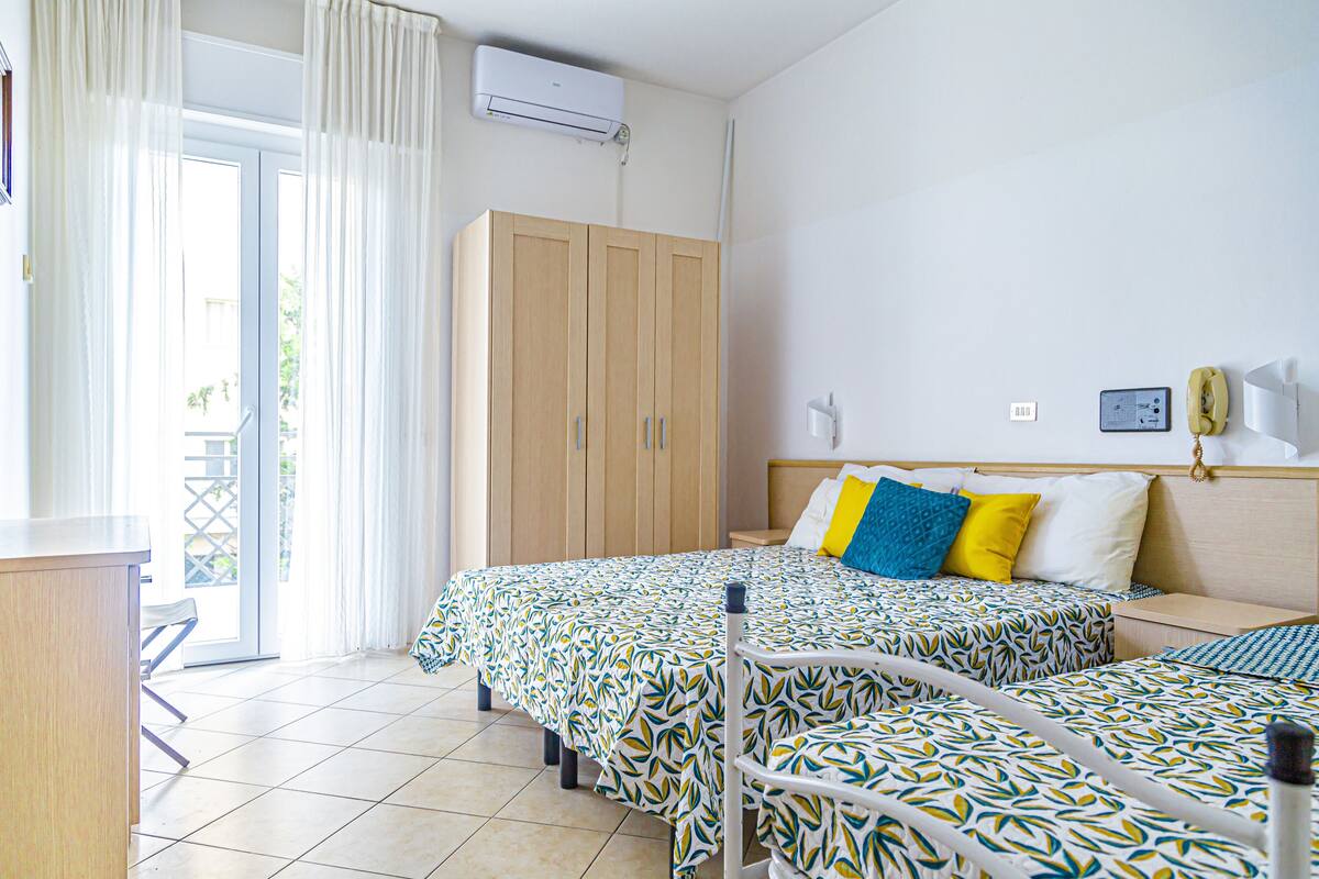 A hotel room with two beds, a wooden wardrobe, a desk, and a balcony with curtains.