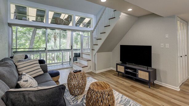 A modern living room with a large TV, comfortable seating, a staircase, and large windows offering a view of trees. - Sunset Rentals | Treetops 6104 (Hilton Head Island)