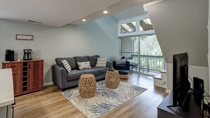 A modern living room with a grey sofa, a coffee station, and a large window offering a view of greenery. - Sunset Rentals | Treetops 6104 (Hilton Head Island)