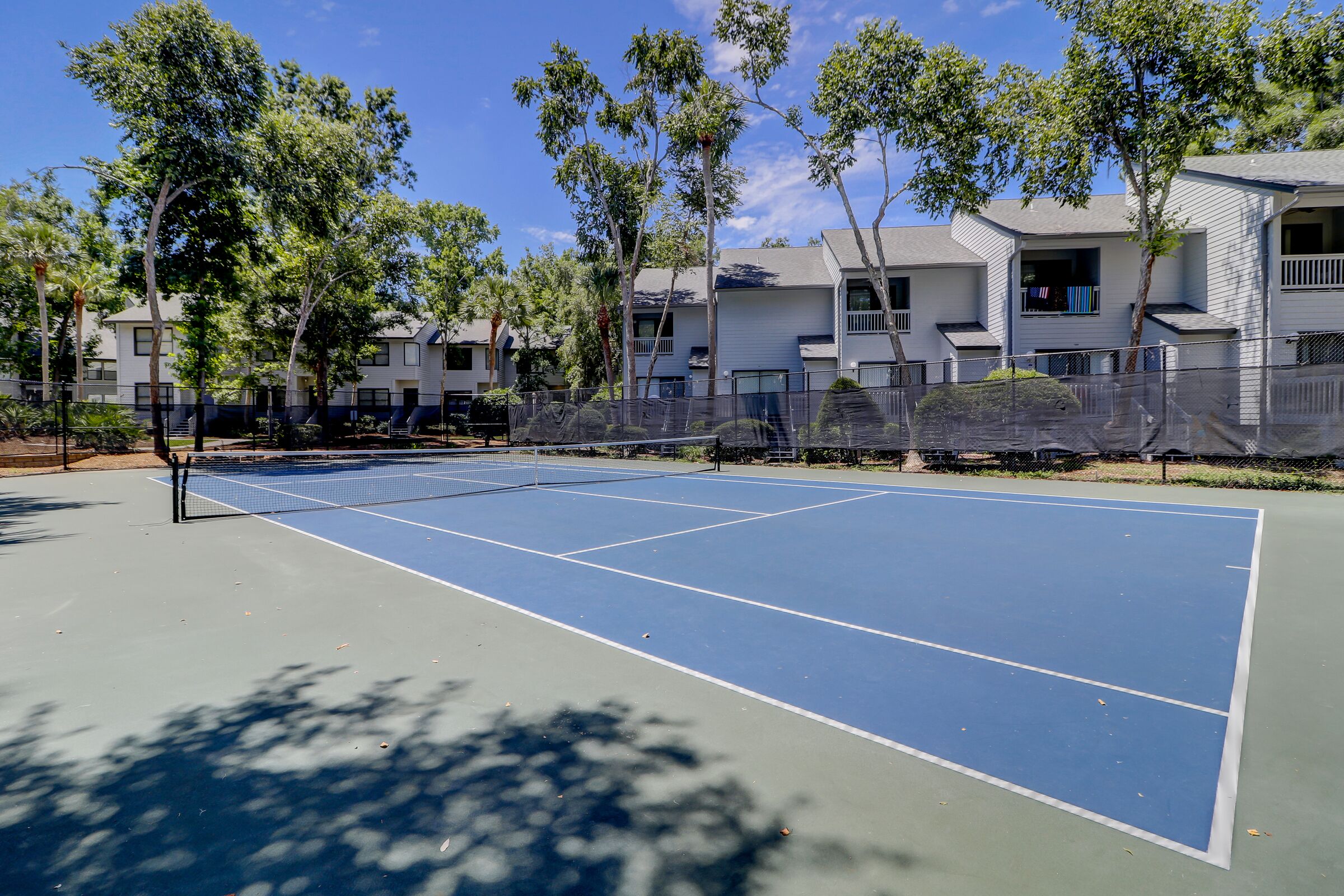 An outdoor tennis court surrounded by trees and a row of white buildings with balconies.