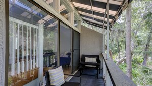 A covered balcony with a view of a forested area, a seating area with a chair and a table, and a sliding glass door leading to an interior space. - Sunset Rentals | Treetops 6104 (Hilton Head Island)