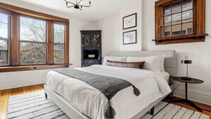 A bedroom with a large bed, a wooden floor, a chandelier, a fireplace, and a window with a view of trees. - Chic Victorian Manor | Sleeps 12 | Congress Park (Denver)