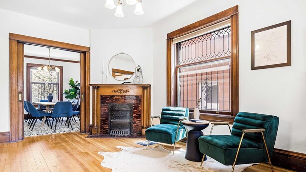 A spacious living room with a fireplace, a dining area with blue chairs, and a large window with a view. - Chic Victorian Manor | Sleeps 12 | Congress Park (Denver)