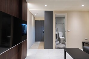 A modern hotel room with a large flat-screen TV mounted on the wall, a small seating area, and a bathroom with a toilet and sink.