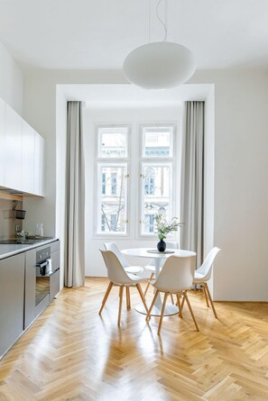 A modern kitchen with white cabinets, a wooden herringbone floor, and a dining area with a round table and four white chairs. - Apartment BRNO (Brno)