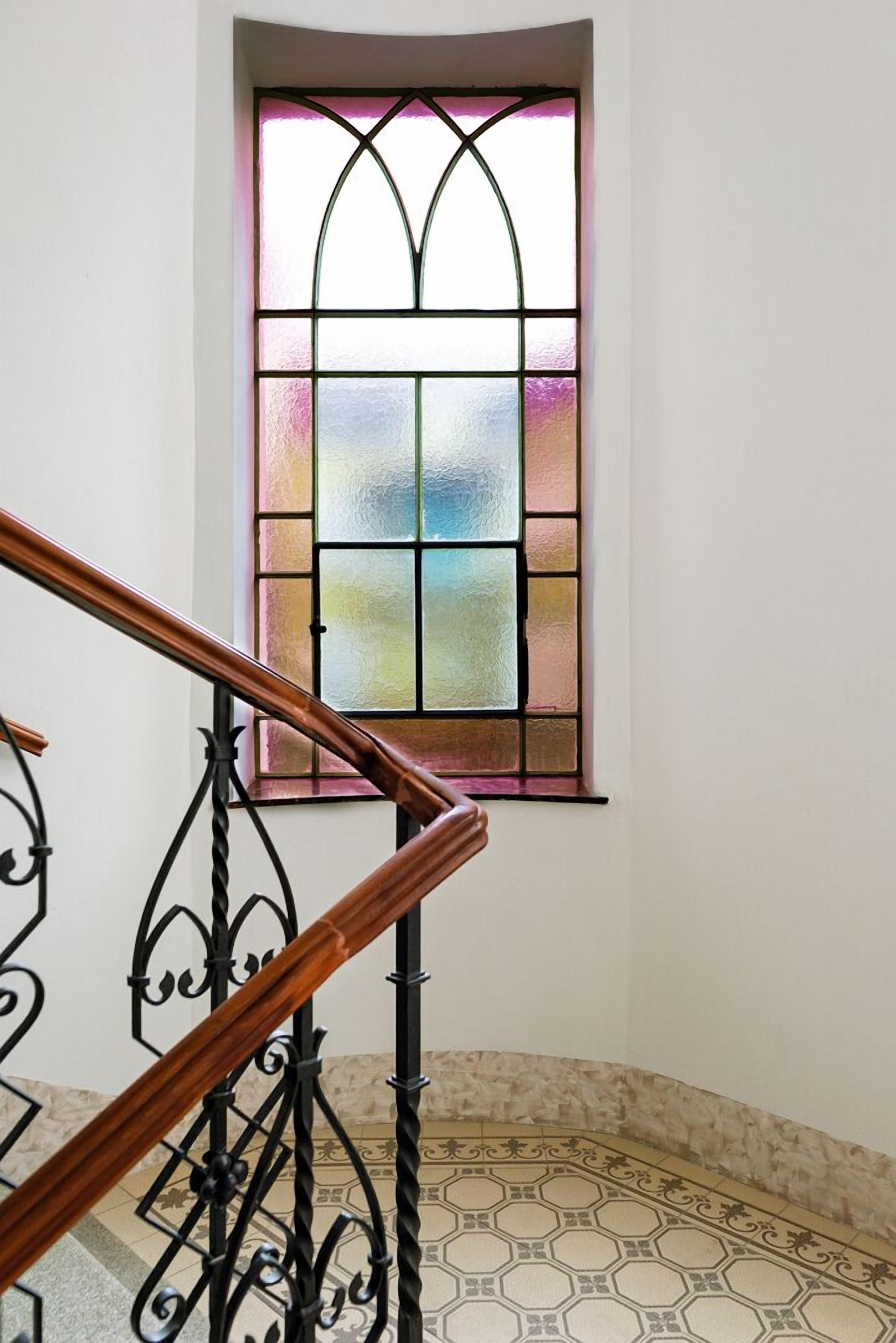 A staircase with a wooden handrail and black metal balusters, leading to a room with a large stained glass window.