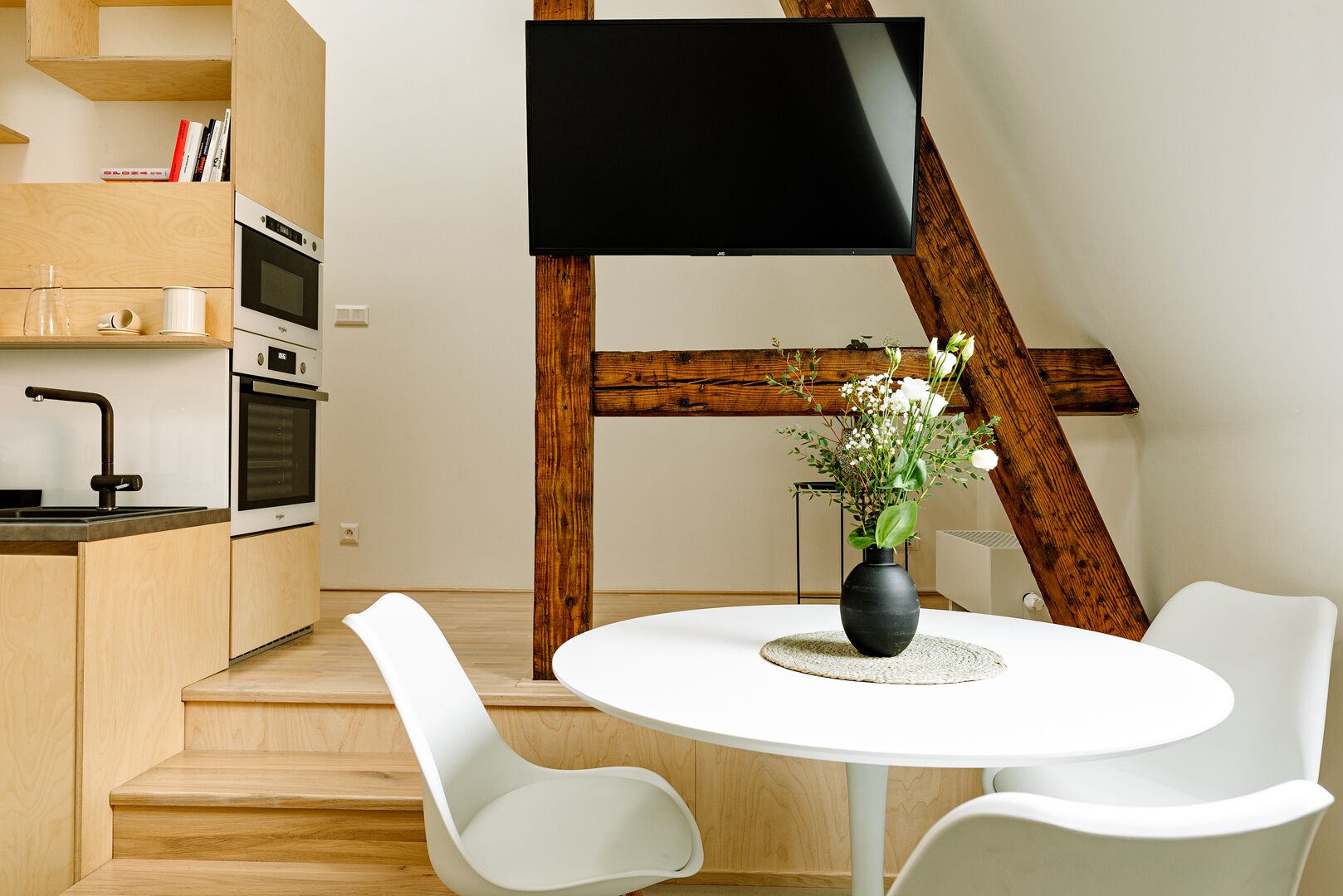 Una cocina moderna con horno empotrado y microondas, una zona de comedor con mesa redonda y sillas blancas, y un televisor de pantalla plana montado en una viga de madera.