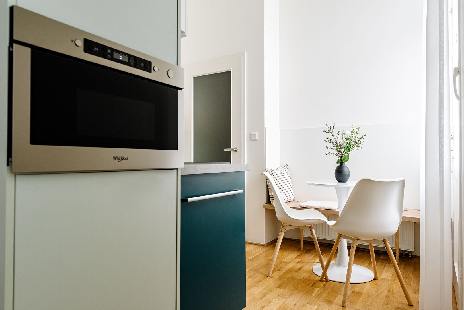A compact kitchen with a built-in oven and a small refrigerator. There is a dining area with two chairs and a table, and a potted plant on the table.