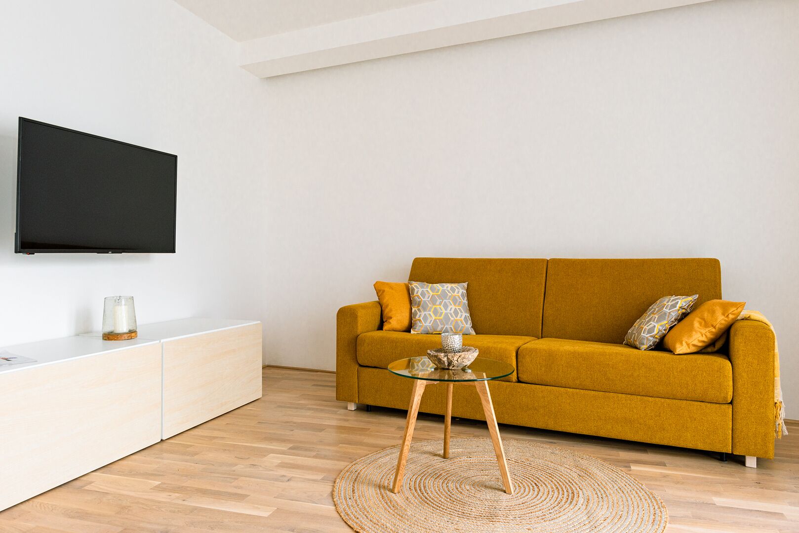 A living room with a mustard sofa, a glass coffee table, a flat-screen TV, and a white cabinet.