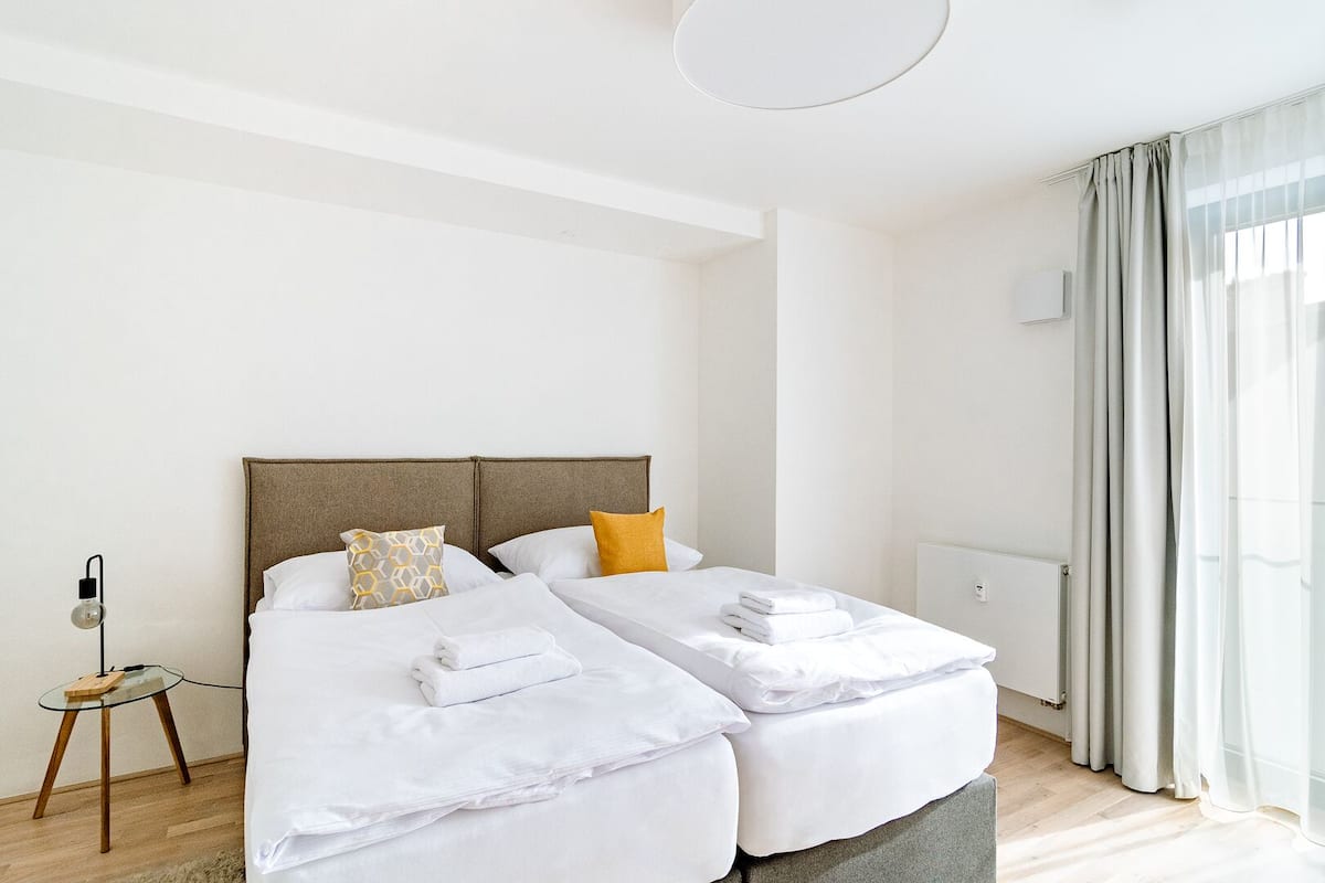 A hotel room with two beds, a small table, a lamp, and a window with curtains.