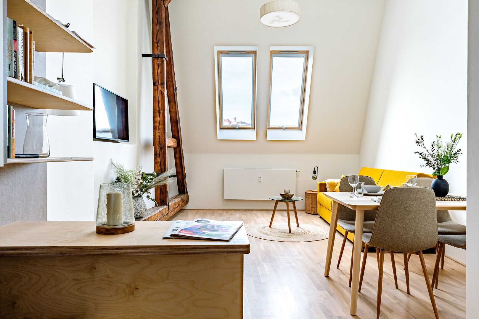 A modern living room with a wooden desk, a yellow sofa, and a dining table with chairs. There are two windows with a view of the sky, a ladder, and a bookshelf with books and decorative items.