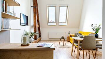 A modern living room with a wooden desk, a yellow sofa, and a dining table with chairs. There are two windows with a view of the sky, a ladder, and a bookshelf with books and decorative items.
