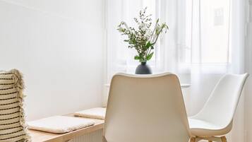A modern room with a wooden floor, a white radiator, a white chair, a white table with a striped cushion, and a window with sheer curtains and a potted plant.