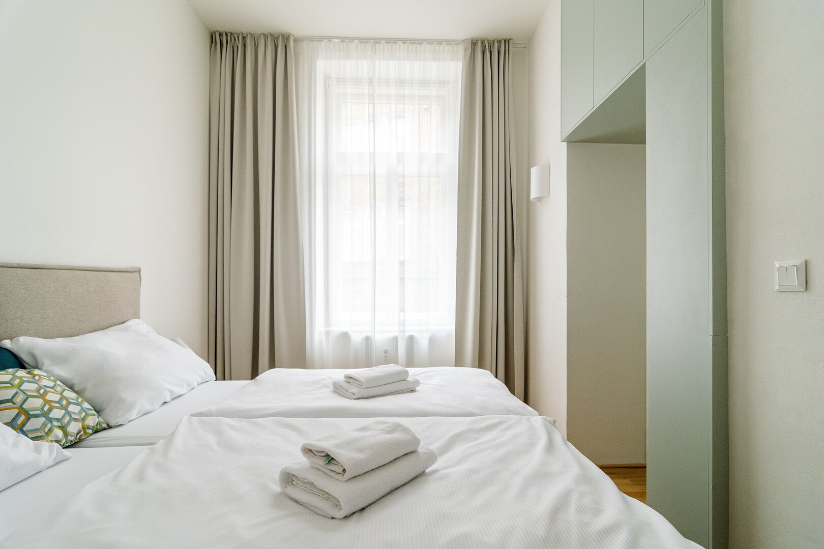 A hotel room with a bed, two pillows, a window with curtains, and a light switch on the wall.