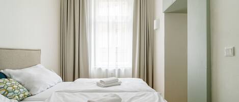 A hotel room with a bed, two pillows, a window with curtains, and a light switch on the wall.