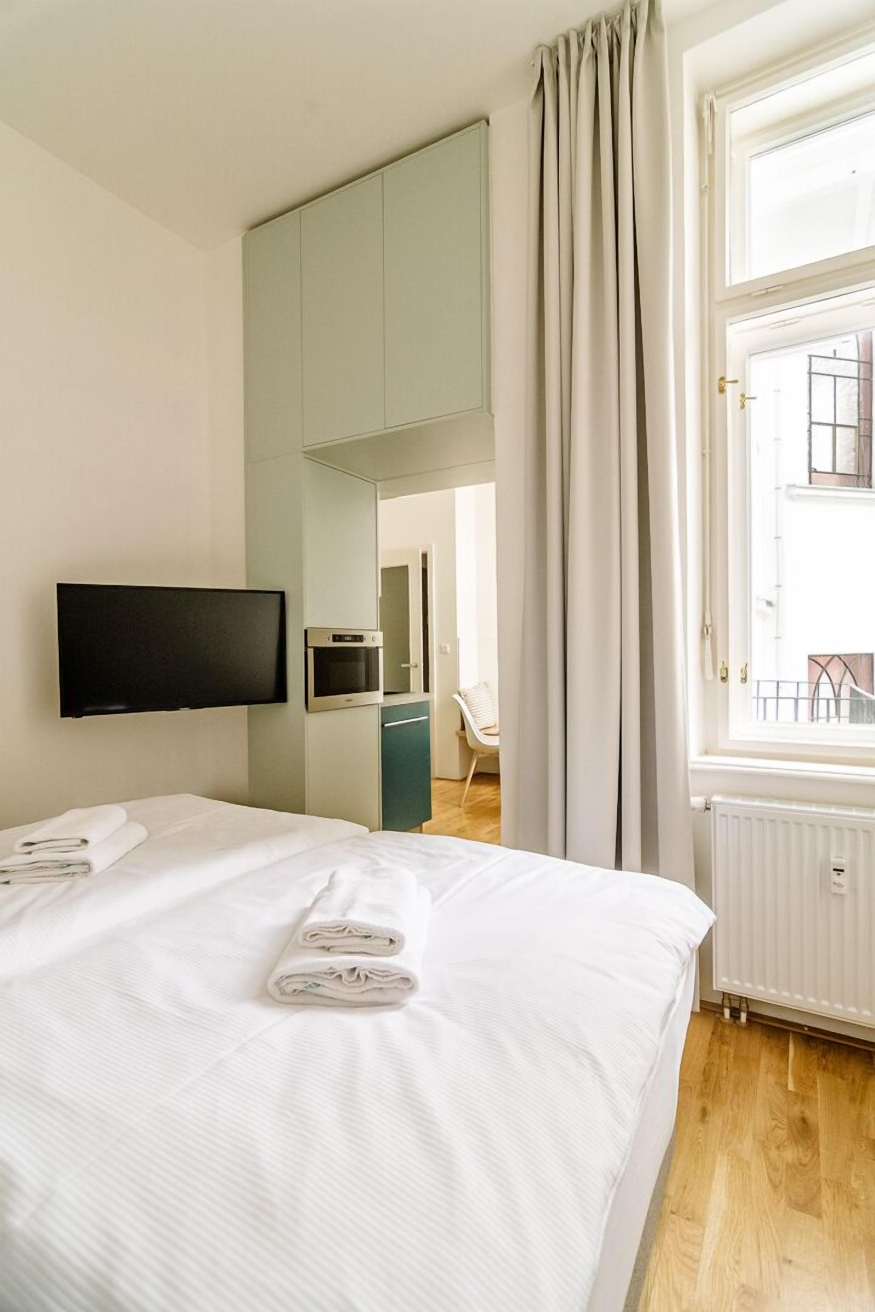 A hotel room with a bed, a flat-screen TV mounted on the wall, a small kitchenette, and a window with curtains.