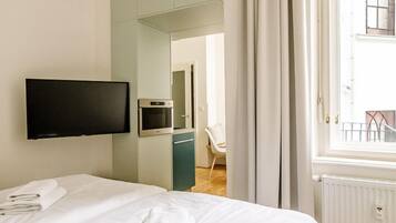 A hotel room with a bed, a flat-screen TV mounted on the wall, a small kitchenette, and a window with curtains.