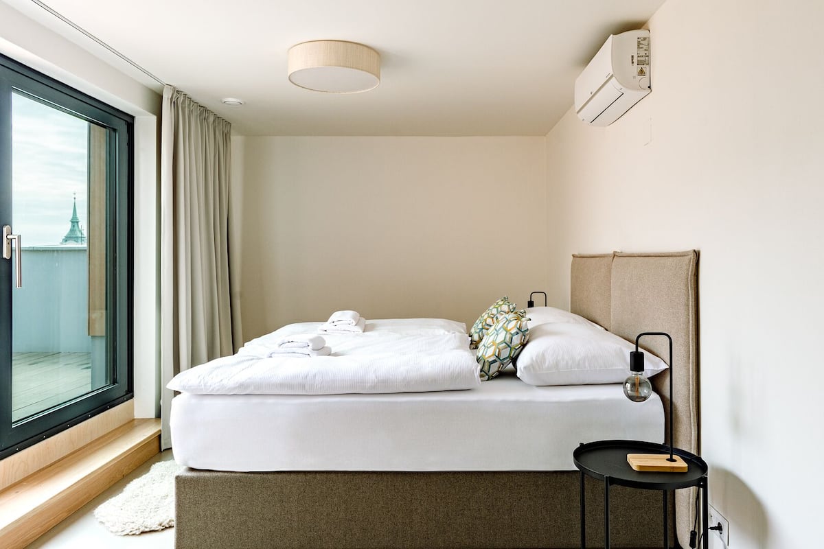 A hotel room with a bed, a bedside table, a lamp, and a view of a cityscape through a large window.