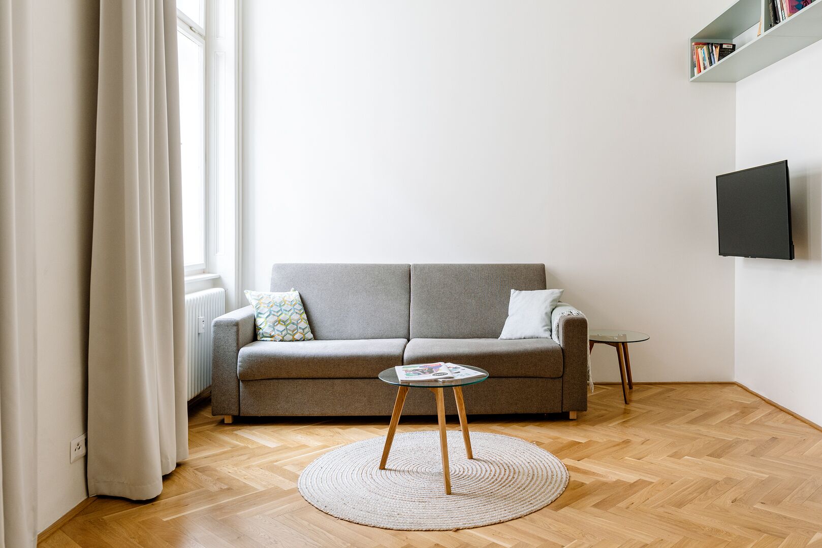 A living room with a grey sofa, a small round table, a TV mounted on the wall, and a bookshelf with books.