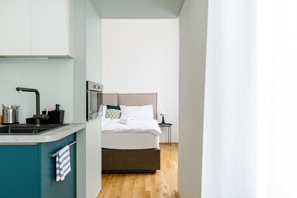 A modern hotel room with a kitchenette, a bed with white linens, and a small bedside table with a lamp. - Apartment BRNO (Brno)