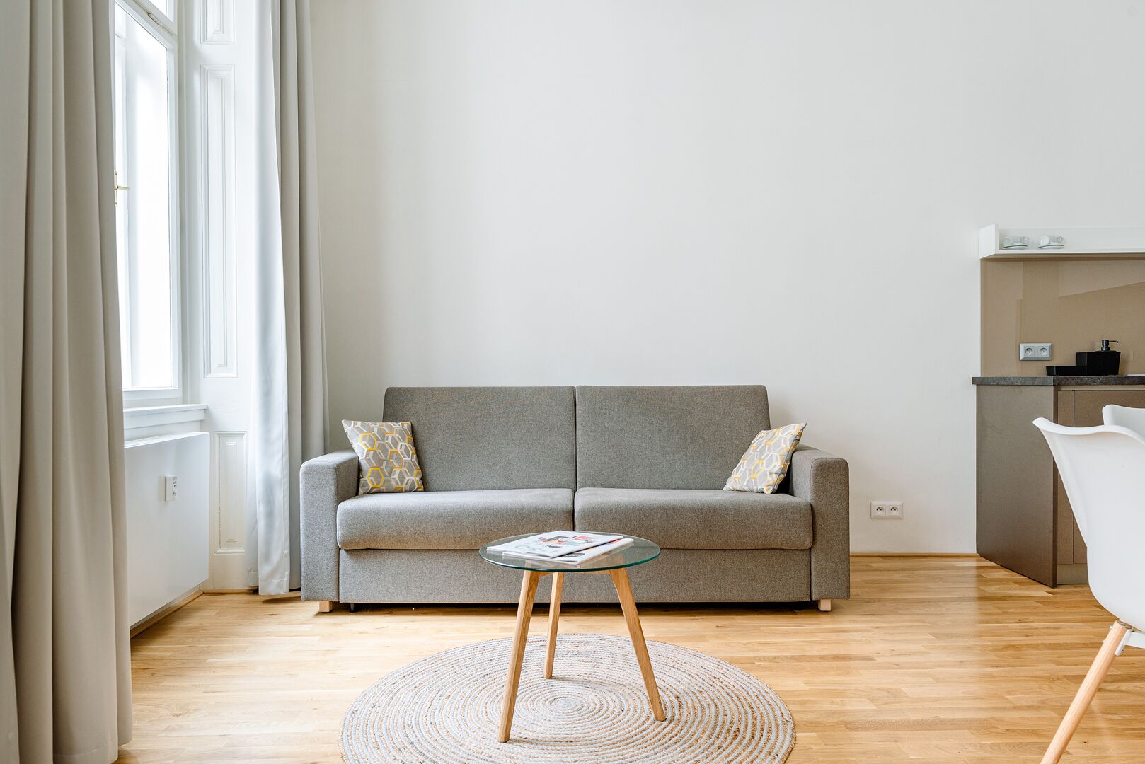 Een moderne woonkamer met een grijze bank, een glazen salontafel en een keuken in de achtergrond.