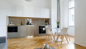 A modern kitchen with white cabinets, a dining area with a white table and chairs, and a large window providing natural light. - Apartment BRNO (Brno)